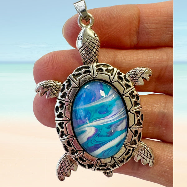turtle shaped pendant with blue stone held in a hand against a beach background