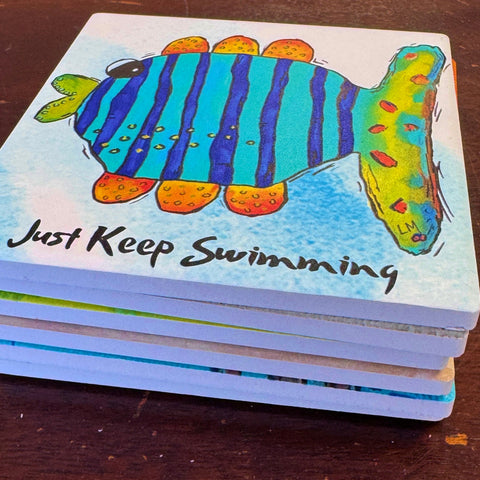 A stack of square coasters on a dark wooden table, featuring a colorful fish illustration and the phrase 'Just Keep Swimming' in black script.