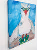 A  canvas print of a white seagull with a red beak, wearing green sunglasses and standing on green kumquats against a bright blue background, shown hanging on a white wall.