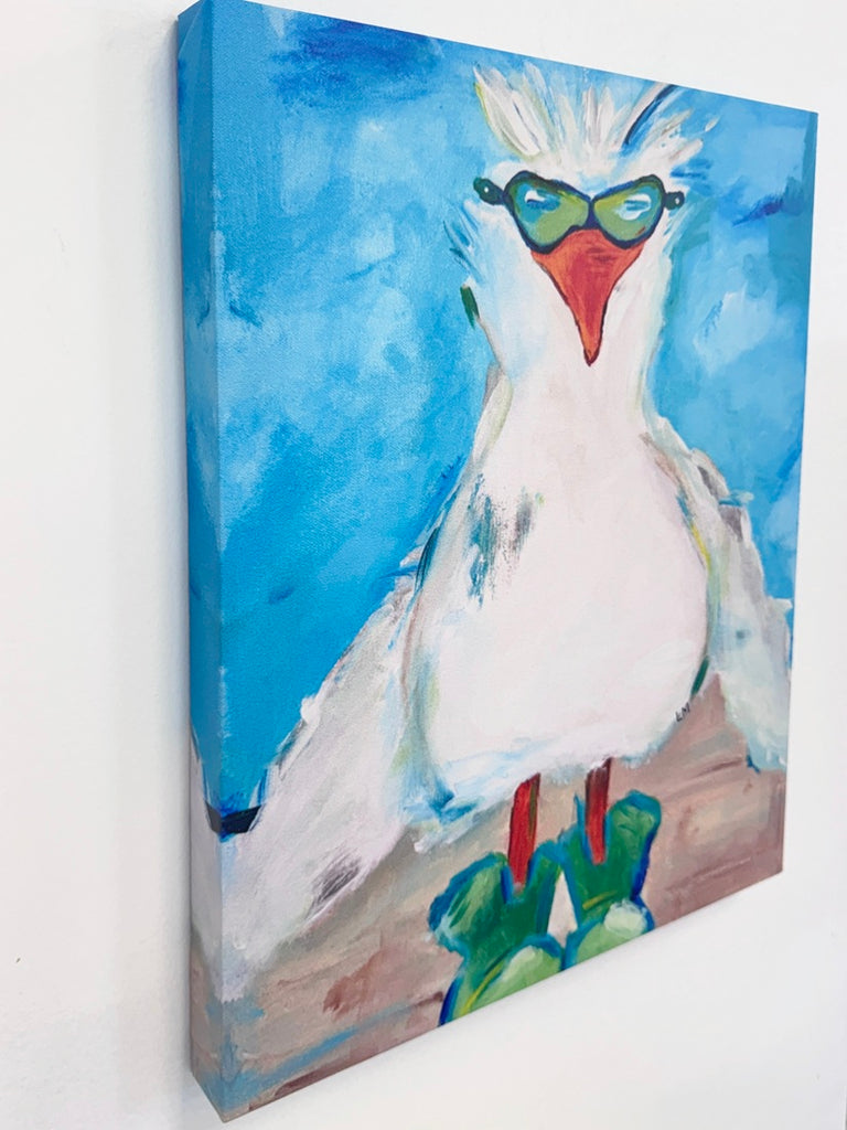 A  canvas print of a white seagull with a red beak, wearing green sunglasses and standing on green kumquats against a bright blue background, shown hanging on a white wall.