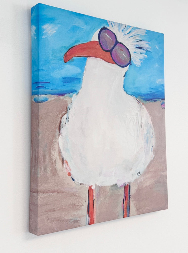 A canvas wall art print showing a stylized white seagull with a red-orange beak, purple-tinted sunglasses, and a blue sky and sandy beach background, photographed hanging on a white wall.