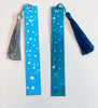 Two painted bookmarks with snow artwork on blue backgrounds.  One with a silver tassel