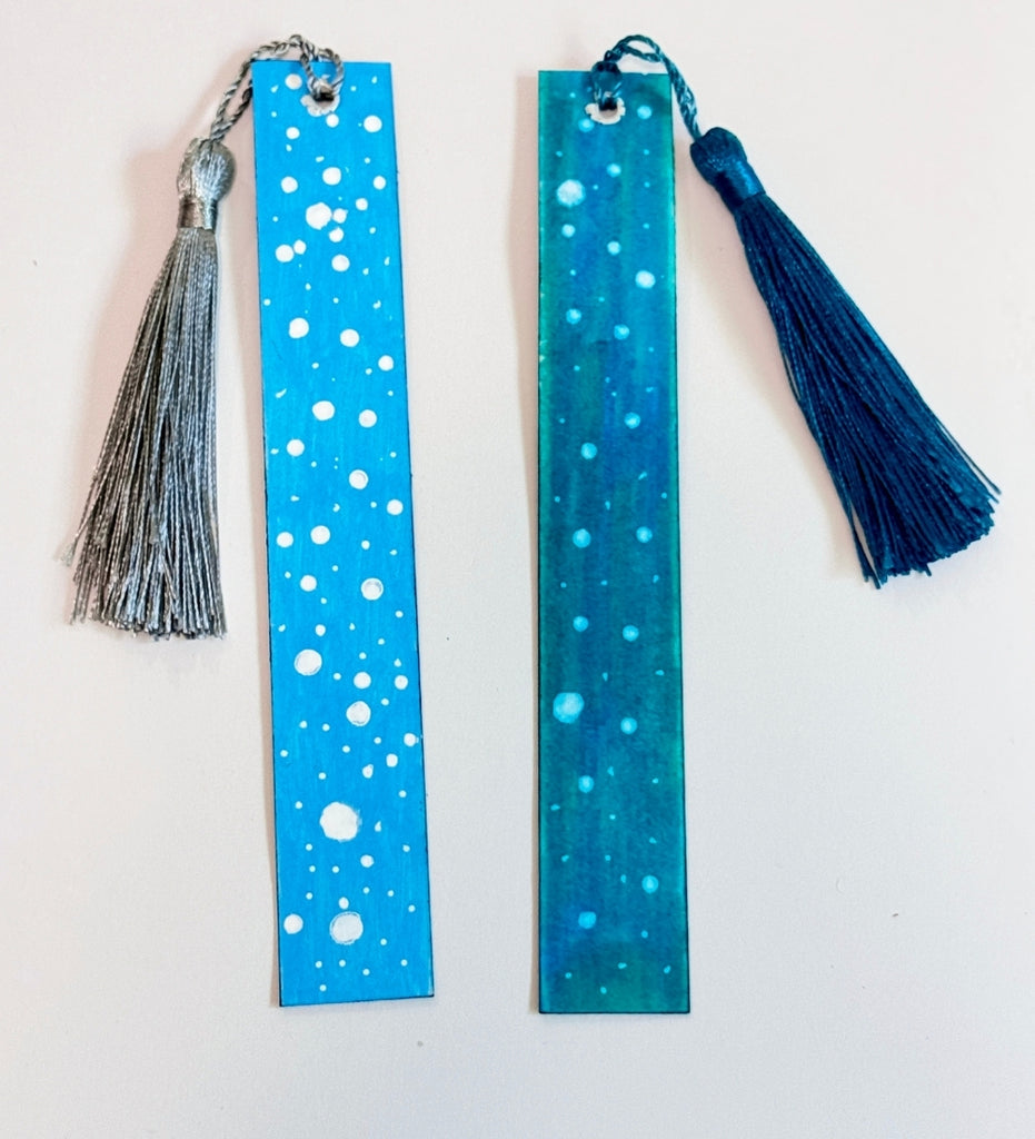 Two painted bookmarks with snow artwork on blue backgrounds.  One with a silver tassel