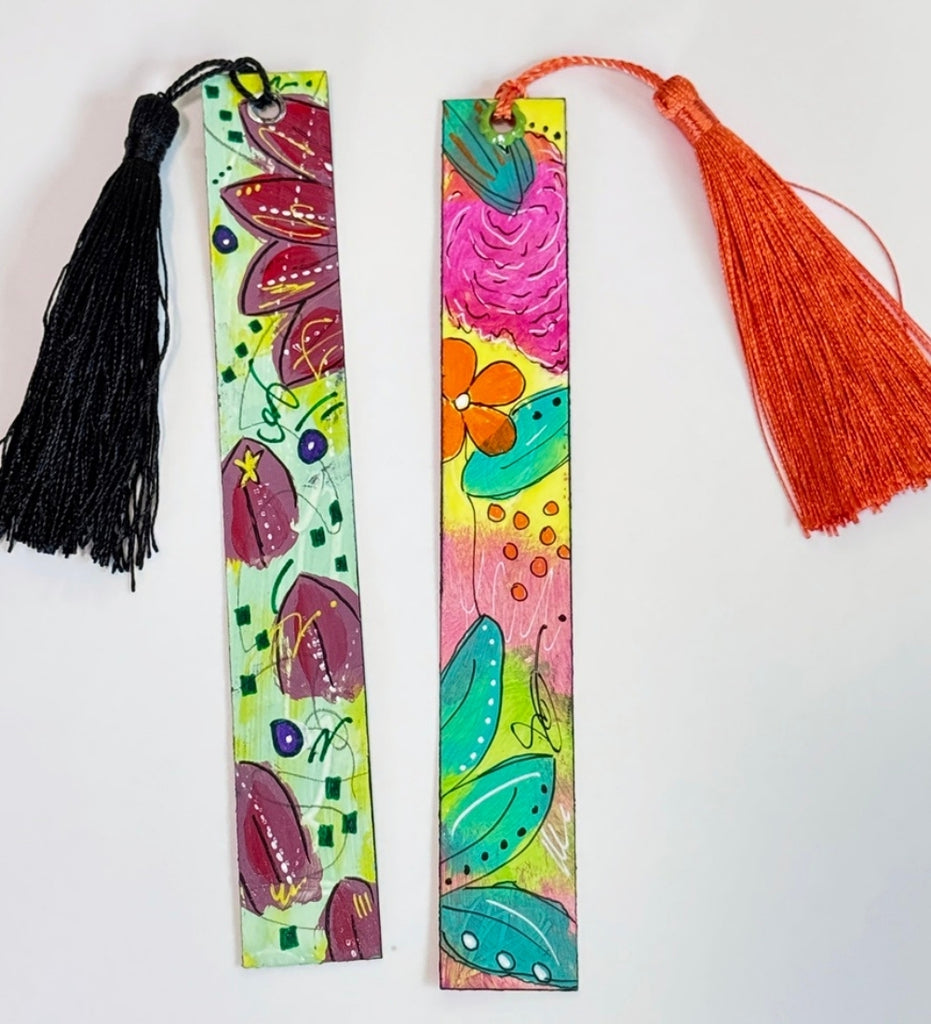 Two colorful hand-painted bookmarks with tassels on a white table. One bookmark has a black tassel and a green base with purple and orange floral designs