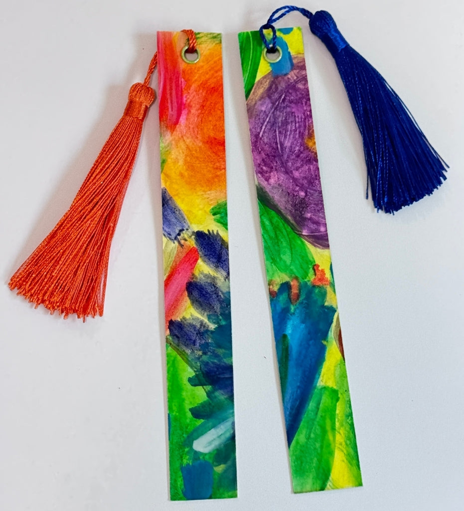 Two colorful bookmarks with tassels on a plain white table. One has a orange tassel and the other has a dark blue tassel.