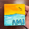 Small painted canvas with a sunset and the word 'AMI' held by a hand on a wooden surface