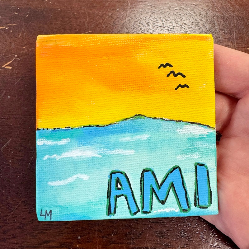 Small painted canvas with a sunset and the word 'AMI' held by a hand on a wooden surface