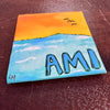 Small painted canvas with a sunset over water scene and 'AMI' text, pictured on a wooden surface.
