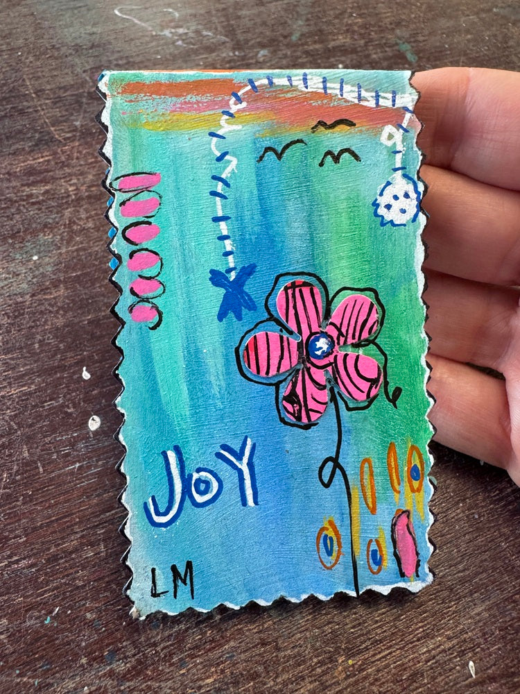 A hand holding a small rectangular handmade art piece with scalloped edges, painted with a blue and green gradient background, a pink flower, and the word 'Joy' in white. The piece is photographed on a dark wooden surface.
