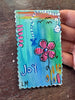 A hand holding a small rectangular handmade art piece with scalloped edges, painted with a blue and green gradient background, a pink flower, and the word 'Joy' in white. The piece is photographed on a dark wooden surface.