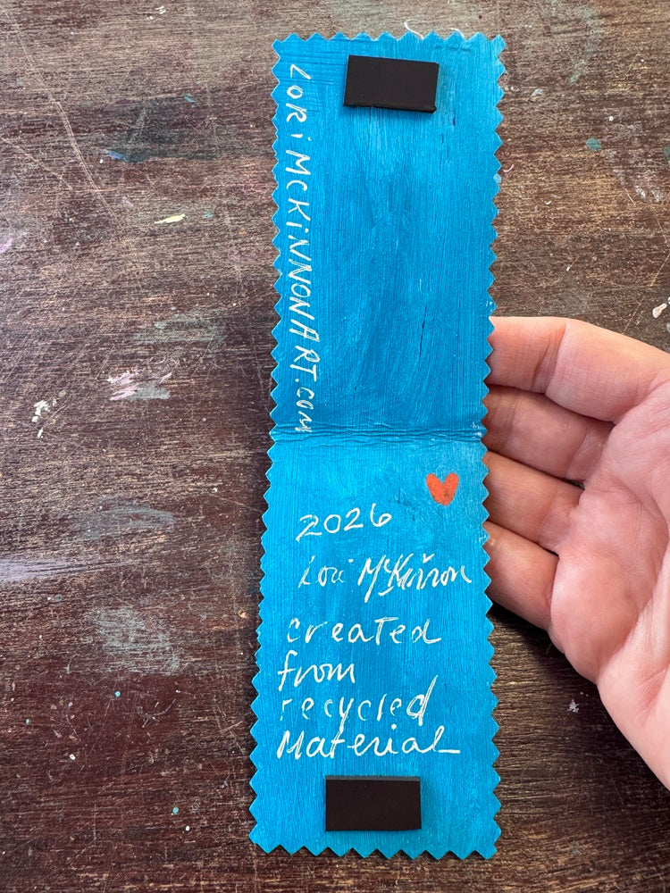 A handmade bookmark with serrated edges, held by a hand over a rustic wooden table. The card features white handwritten text, a small orange heart, and two black rectangular magnets near the top and bottom.