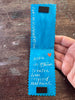 A handmade bookmark with serrated edges, held by a hand over a rustic wooden table. The card features white handwritten text, a small orange heart, and two black rectangular magnets near the top and bottom.