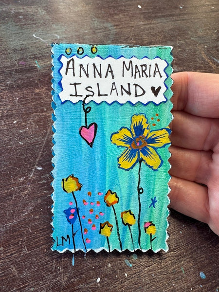 A handmade rectangular magnetic bookmark with scalloped edges, featuring the text 'ANNA MARIA ISLAND' and colorful painted flowers and hearts on a blue background, held by a hand over a dark wooden surface.