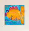 A square canvas painting featuring a whimsical orange and yellow fish with blue eyes on a turquoise background, with the handwritten text 'Don't take the bait' in blue. The canvas is mounted on a light beige wall.