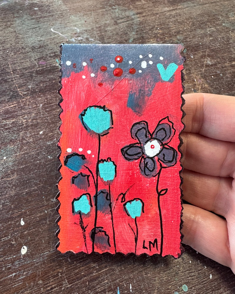 A small hand-painted bookmark with a red background, black scalloped edges, and stylized flowers in teal, black, and white. A hand is holding the item on a dark wooden surface.