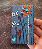 A hand holds a rectangular painting with gray background. The bookmark features hand-painted orange flowers, black stems, white script text reading 'BE you', and small turquoise accents.