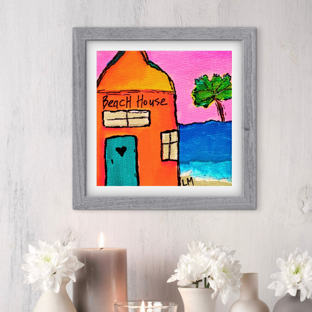 Framed artwork of a beach house with a palm tree on a pink sky, displayed on a wall with decorative elements.