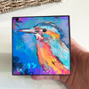 Birds of Color art block held in one hand for scale, showing a turquoise, orange, and pink bird on a blue background 