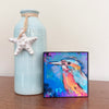 Birds of Color art block featuring a turquoise, orange, and pink bird, displayed on a wooden tabletop beside a pale blue vase with a white starfish ornament.