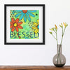 Framed artwork with colorful flowers and the word 'Blessed' next to a vase with flowers on a wooden surface.