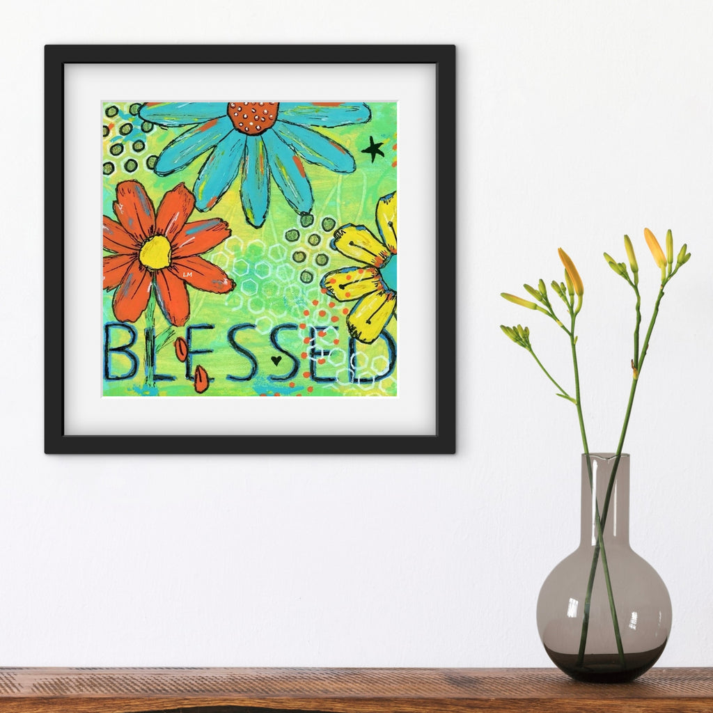Framed artwork with colorful flowers and the word 'Blessed' next to a vase with flowers on a wooden surface.
