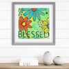 Framed artwork with colorful flowers and the word 'Blessed' on a white wall.