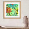 Framed artwork with colorful flowers and 'BLESSED' text on a wall above a wooden shelf.