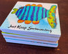 A stack of square coasters on a dark wooden table, featuring a colorful fish illustration and the phrase 'Just Keep Swimming' in black script.