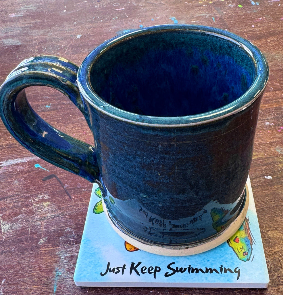 a coffee mug sitting on a square coaster that has text "Just Keep Swimming on a dark tabletop