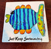 A square printed coaster featuring a colorful fish with blue vertical stripes, orange and yellow accents, and the text 'Just Keep Swimming' in black script, photographed on a dark wooden surface.