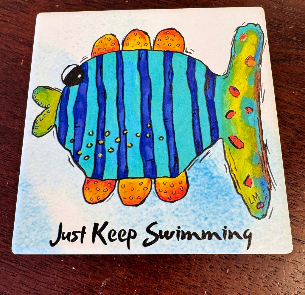 A square printed coaster featuring a colorful fish with blue vertical stripes, orange and yellow accents, and the text 'Just Keep Swimming' in black script, photographed on a dark wooden surface.