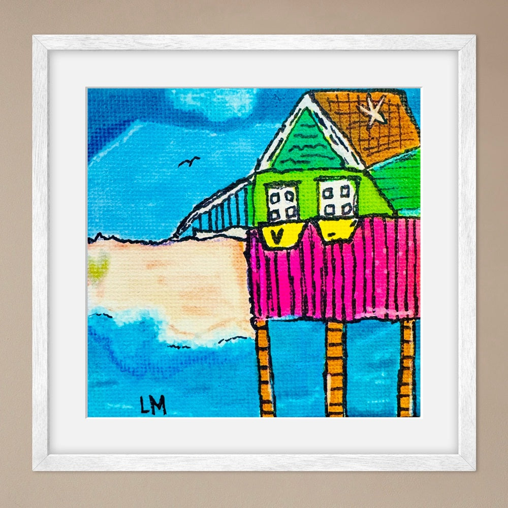 Colorful artwork of a house by the sea with a white frame on a beige wall.