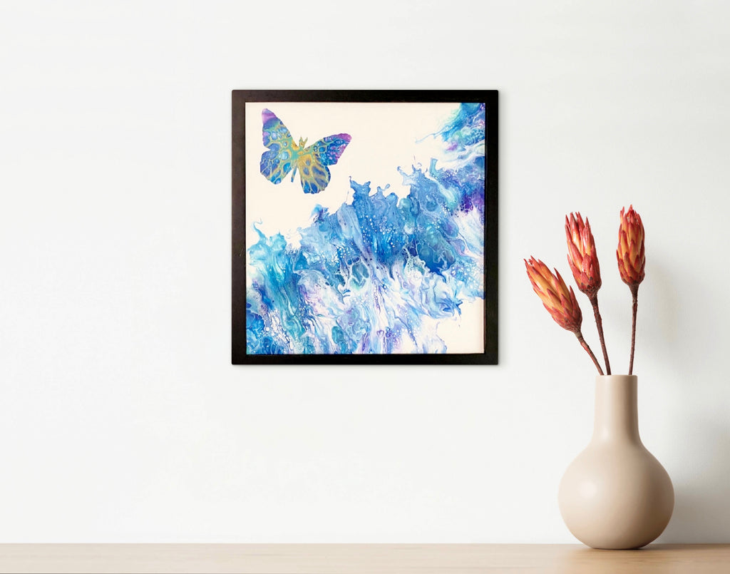 Expansion butterfly painting in a black frame on a white wall beside a cream vase with red-orange flowers