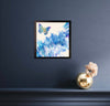 Expansion butterfly painting in a black frame on a dark blue wall above a ledge with a gold vase and pale flowers