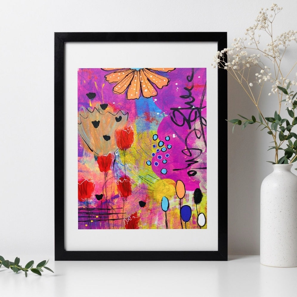 Framed colorful abstract art piece on a white wall with a vase and plant.