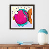 Framed artwork of a colorful fish on a wall above a wooden surface with a vase and books.