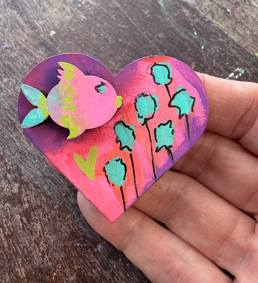 Heart-shaped colorful magnet with a fish and flowers design held by a hand on a wooden surface.