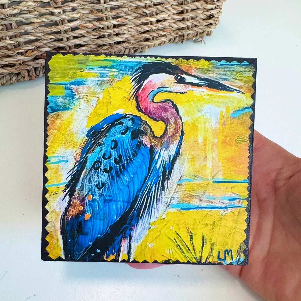 blue heron painting held by a hand over a white table with a basket behind it
