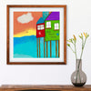 colorful beach home artwork in brown frame on white wall with vase of flowers below
