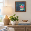 a framed painting on a wall above a table with some books, a plant and a lamp