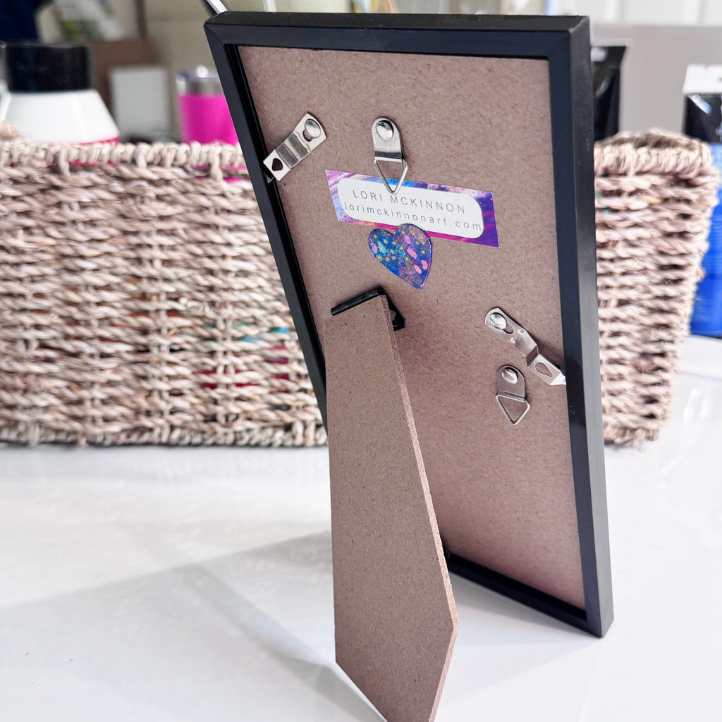 Back of the black frame for the “Pride” art print, showing the easel stand, wall-hanging hardware, and Lori McKinnon Art label