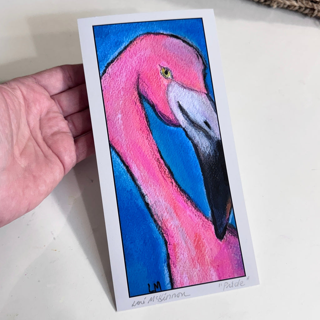 Hand holding the unframed “Pride” art print, showing a pink flamingo against a blue background with a white border, artist signature, and handwritten title.