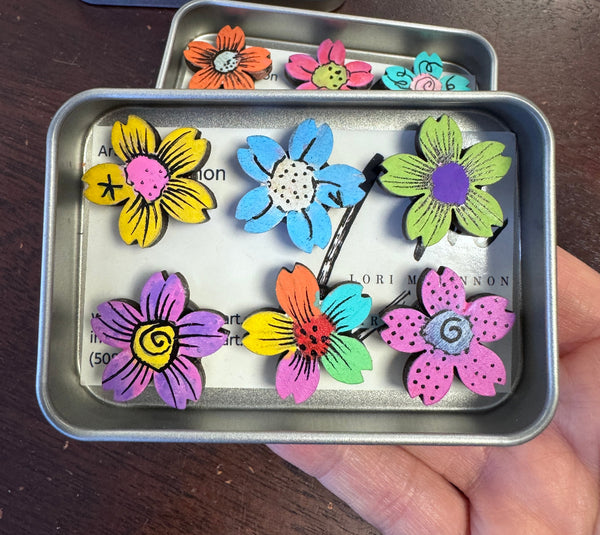 painted flower magnets in a tin being held by a hand above a wooden surface
