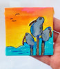 Hand holding a small painted canvas with birds and a sunset scene.