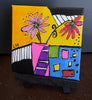 A small square canvas painting displayed on a black easel stand, featuring a colorful abstract floral design with pink flowers, a yellow background, black and white striped shapes, and blocks of blue, green, pink, purple, and red.