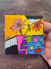A hand holding a small square canvas painting with a colorful abstract floral design on a worn wooden tabletop.