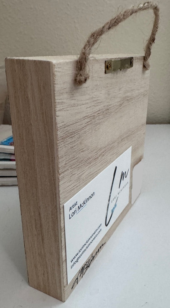 The back of a wooden wall-hanging with a natural light wood grain finish, a rustic rope handle. The label includes the text 'Artist Lori McKinnon' and artist signature