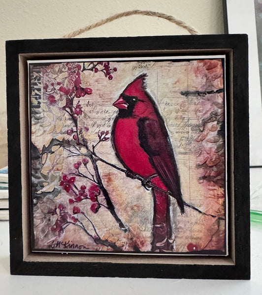 A square framed art print featuring a red cardinal perched on a branch with red berries, set against a vintage-style beige background with script text. The piece is housed in a dark wood frame with a hanging twine loop at the top.