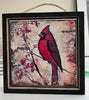 A square framed art print featuring a red cardinal perched on a branch with red berries, set against a vintage-style beige background with script text. The piece is housed in a dark wood frame with a hanging twine loop at the top.