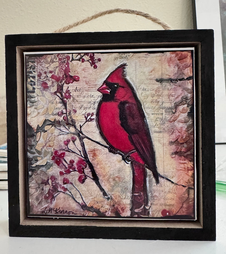 A square framed art print featuring a red cardinal perched on a branch with red berries, set against a vintage-style beige background with script text. The piece is housed in a dark wood frame with a hanging twine loop at the top.